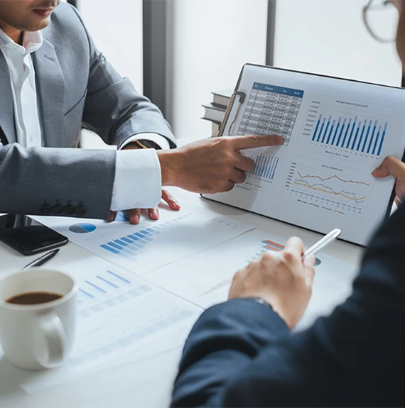 A professional business setting where two people in suits are reviewing financial reports. One person points to a data table on a clipboard while the other holds a pen, illustrating expert advice & strategic guidance through a detailed analysis of bar charts and line graphs.