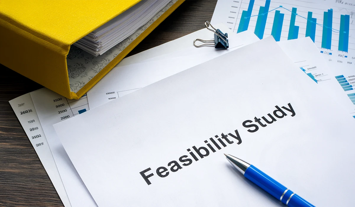 A professional desk setup featuring a document titled "Feasibility Study" in bold text, accompanied by a blue pen, a yellow binder, and various financial bar charts and data sheets used by feasibility study companies in UAE to evaluate project viability.